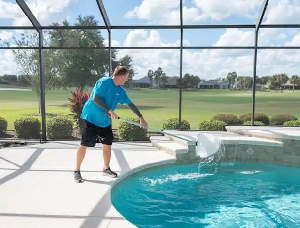 A man in a blue shirt uses a pool skimmer to clean debris from a backyard swimming pool on a sunny day. The sparkling pool, serviced by Patriot Pool and Spa, is enclosed with screens and overlooks a green lawn and trees.