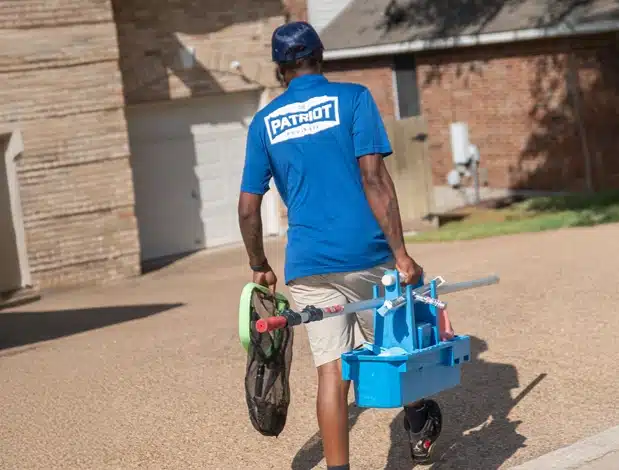 A person in a blue "Patriot Pool and Spa" t-shirt and khaki shorts walks on a driveway, carrying pool cleaning equipment and a net. The surroundings include a brick building and a house.