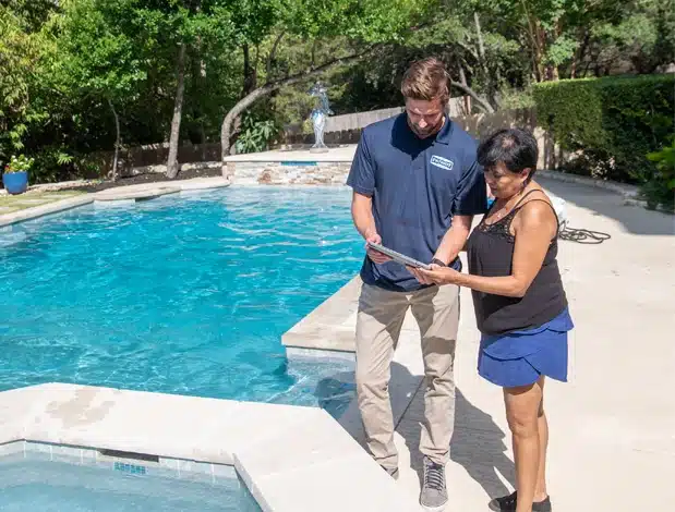 A man and woman stand by a backyard pool and hot tub on a sunny day, looking at a clipboard together. The concrete pool area, surrounded by trees and bushes, showcases the quality service of Patriot Pool and Spa.